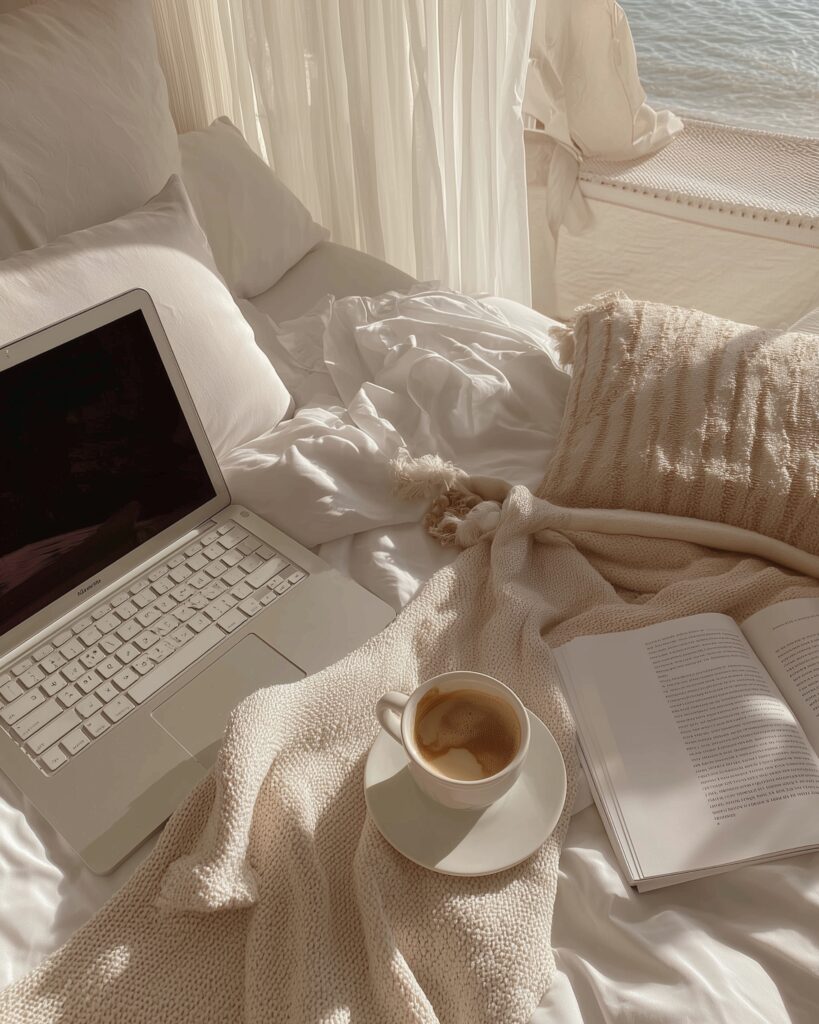 Cozy coastal-inspired bed scene with laptop, coffee, and open book representing reflective writing, creativity, and slow living